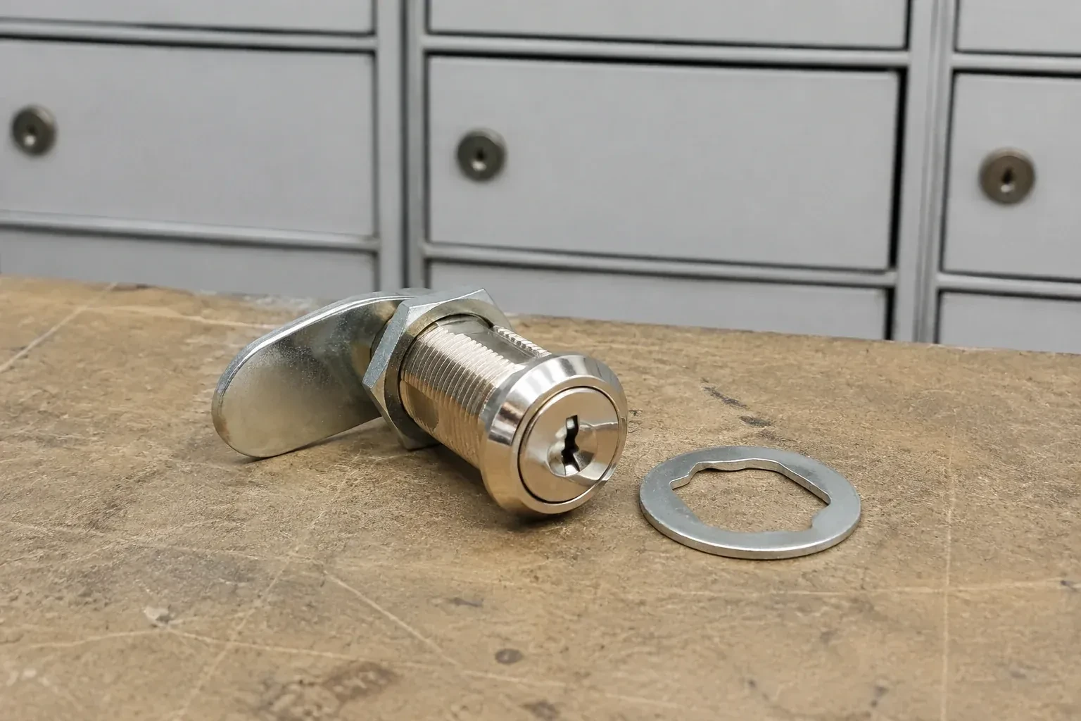 Detached threaded cam lock and mounting washer on a workbench in front of closed mail compartments. — Mailbox Lock Replacement, Markham